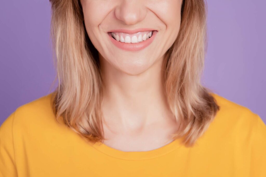 02 Woman with shoulder-length blonde hair smiling in orange T-shirt on mauve background_Lumineers vs. porcelain veneers