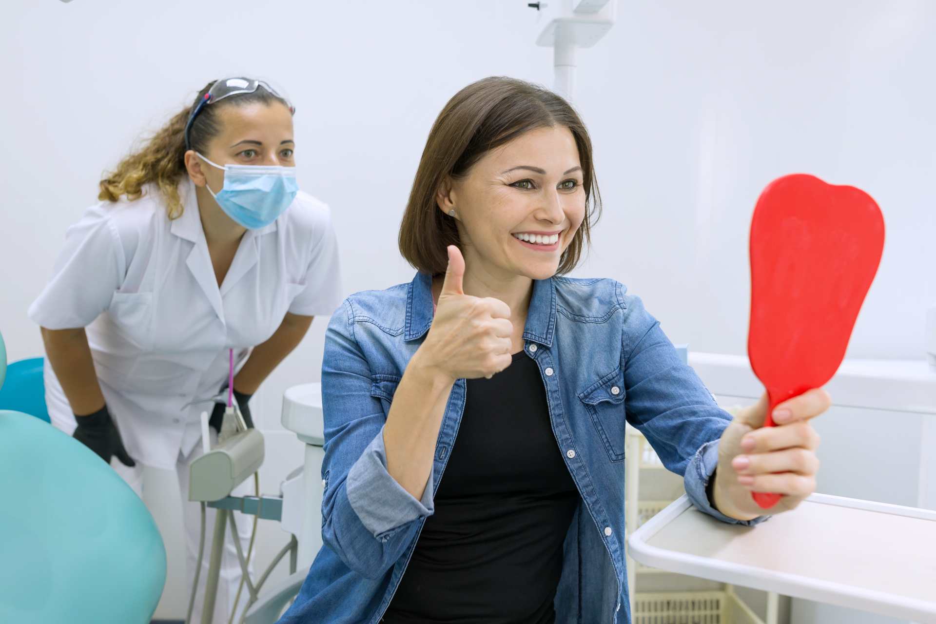 01 Mature woman examining her dental implants with pleasure in front of the mirror next to the dentist_Dental implants pros and cons, dental implants in Miami